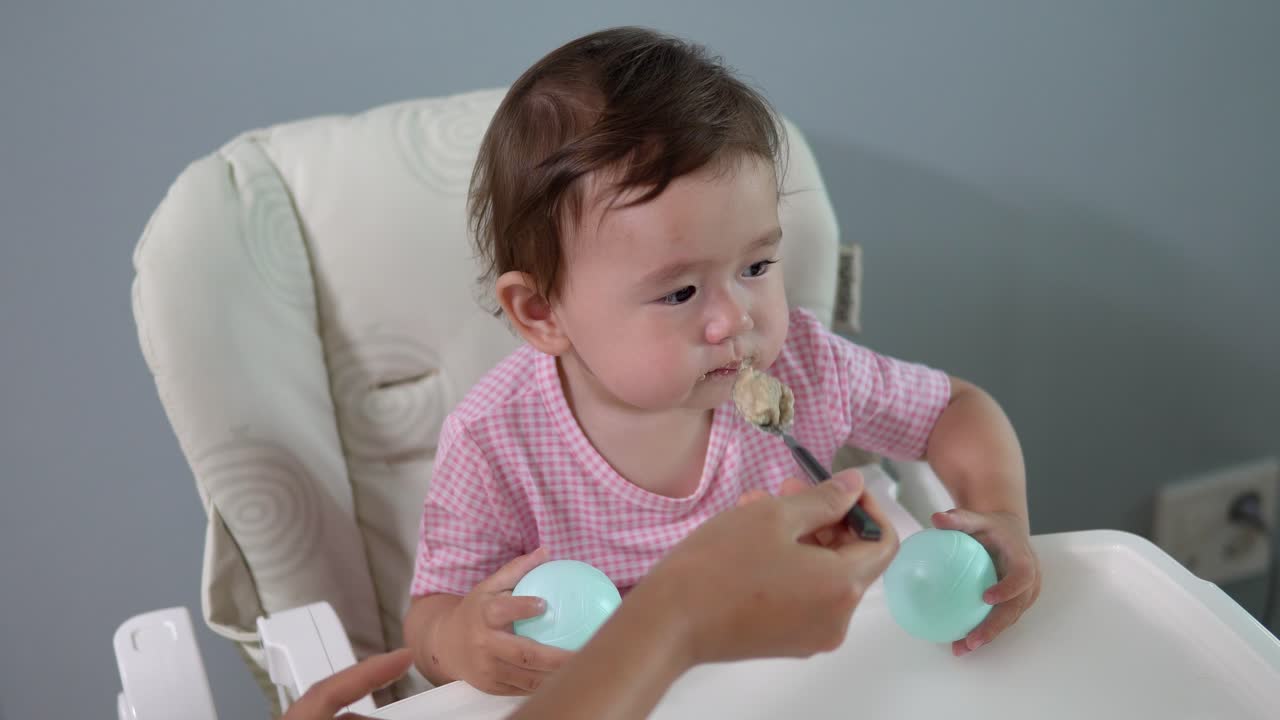 사랑스러운 아시아 유아가 산만하고 지루하며 관심이없는 것처럼 보이는 동안 유아용 의자에서 아기에게 먹이를줍니다.