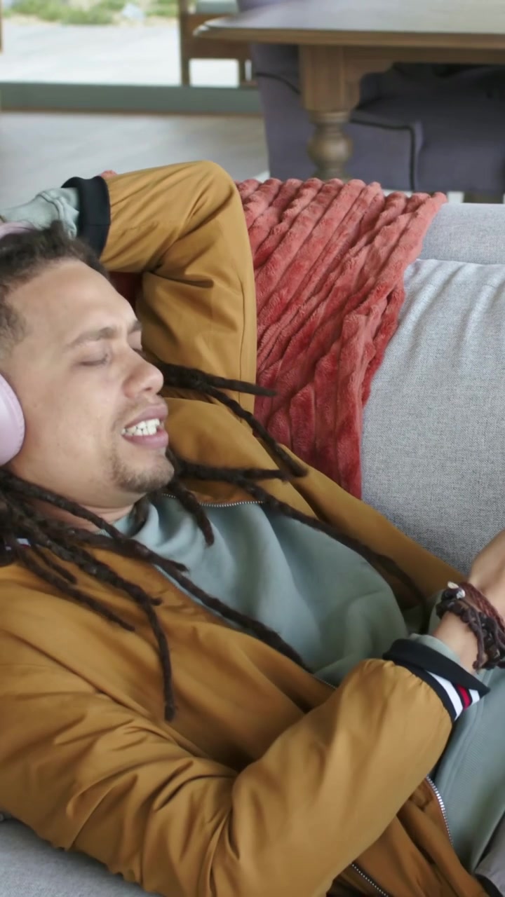Video verticale: Uomo che tocca il telefono, indossa cuffie rosa sul divano, si muove a ritmo di musica e poi scorre.
