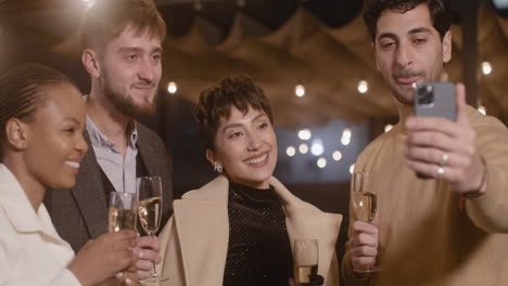 portrait of group of four happy multiethnic friends holding champagne glasses and taking a selfie video at new year's eve party