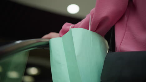 hand view of white lady in pink dress holding mint green shopping bag with a black bag, both hands resting on the escalator rail as she moves up, blurred lights are visible in the background