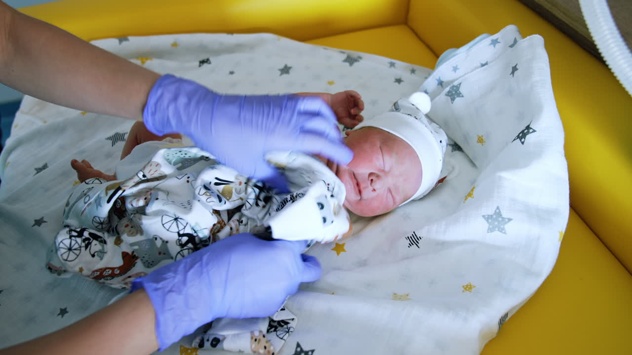 Adorable newborn child is dressed into a romper. Baby with closed eyes doesn't like the procedure.