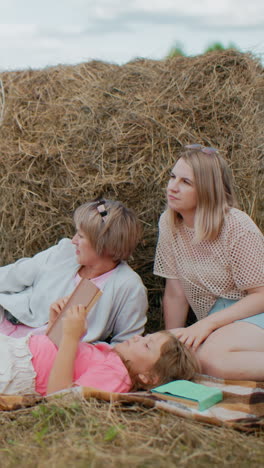 Family retreat with dog on leash, little girl reading book, green book nearby, mother and grandmother relaxing against hay bale in vast countryside