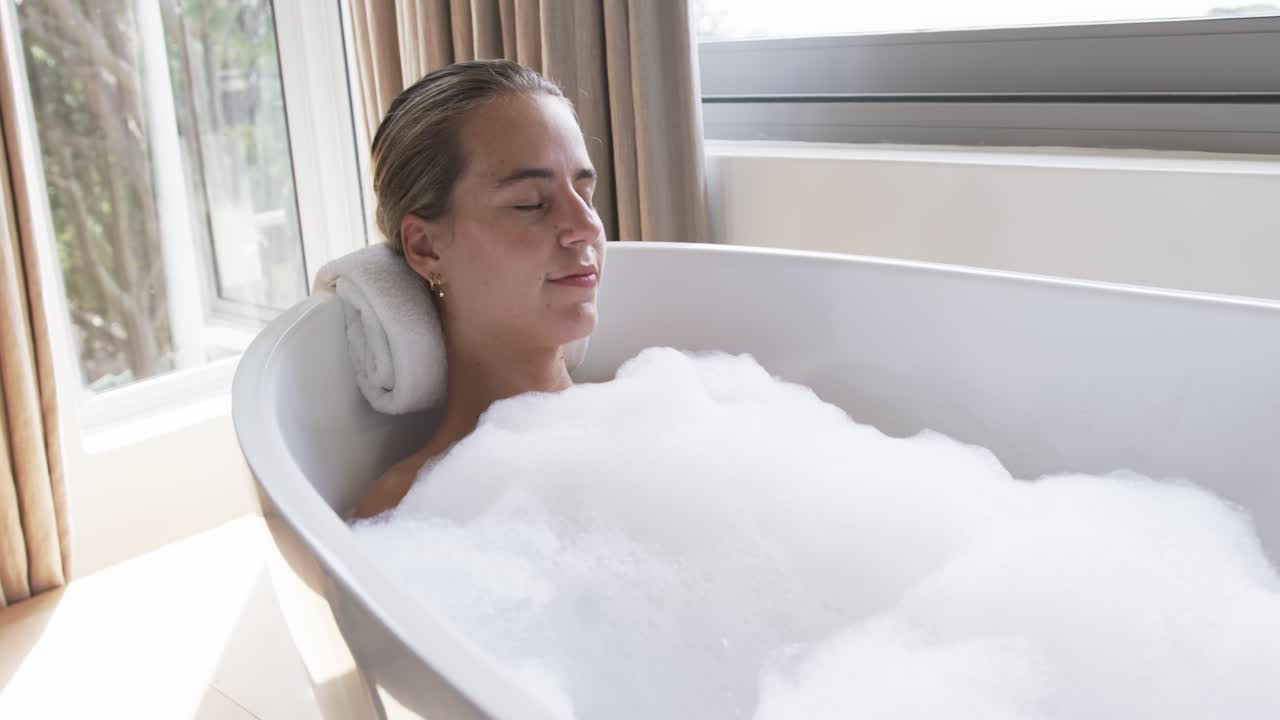 Dopo il bagno di schiuma, una donna sui vent'anni è sdraiata su un asciugamano arrotolato nella vasca, sorridente in segno di relax.
