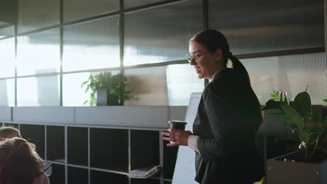 Brunette-businesswoman-in-business-clothes-holds-a-glass-of-coffee-in-her-hands-and-tells-her-colleagues-about-her-ideas-using-a-stand-in-a-sunny-office-during-a-meeting