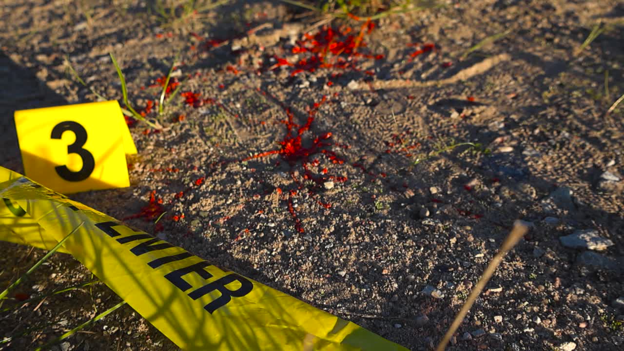 Close up video gliding over a abandoned on unsolved crime area with yellow colored crime scene tape on dirt ground with blood spatter and a crime scene police marker next to it. Sunny day, rural area