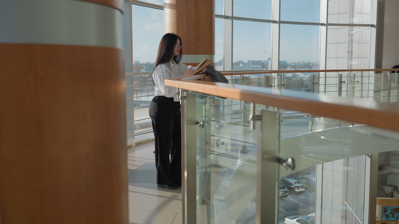 Young girl with long dark hair reads book while resting on rail in modern glass building, with soft natural light and urban scenery outside, including cars moving in street reflections below