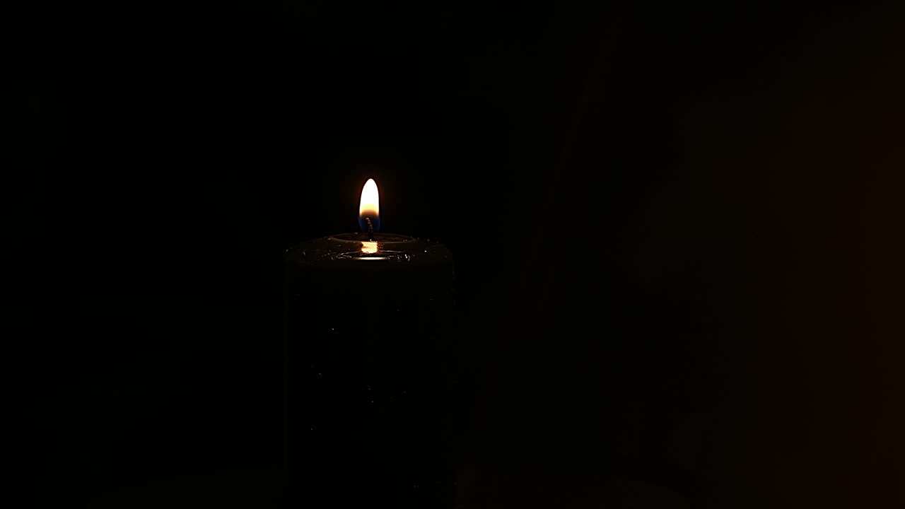 Candela nera accesa in primo piano, volute su sfondo nero con bokeh dorato. Messa a fuoco selettiva.