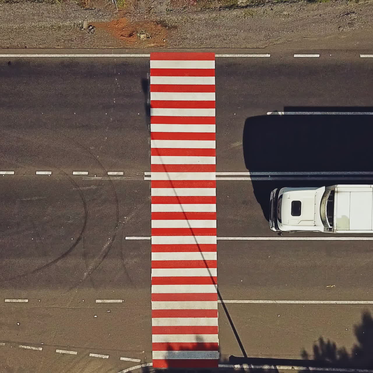 White truck and some cars passing the pedestrian crossing painted with red and white stripes on the road. Zebra crossing on asphalt and cars. Aerial view