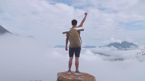 asian hiker male standing on the rock and raising his hand celebrating reaching up top of foggy mountain