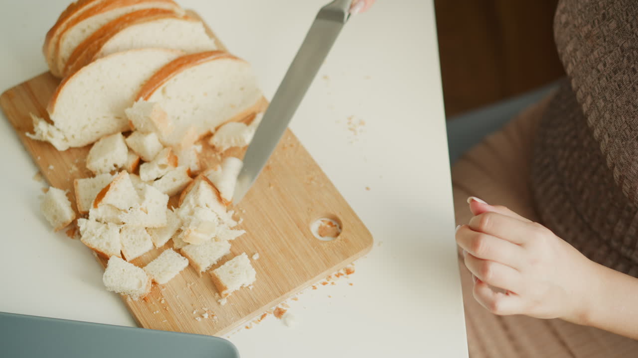 High angle view of baker chopping bread slices into precise small cubes on wooden board over white table, crumbs scattered under soft window light, focused on calm culinary preparation process