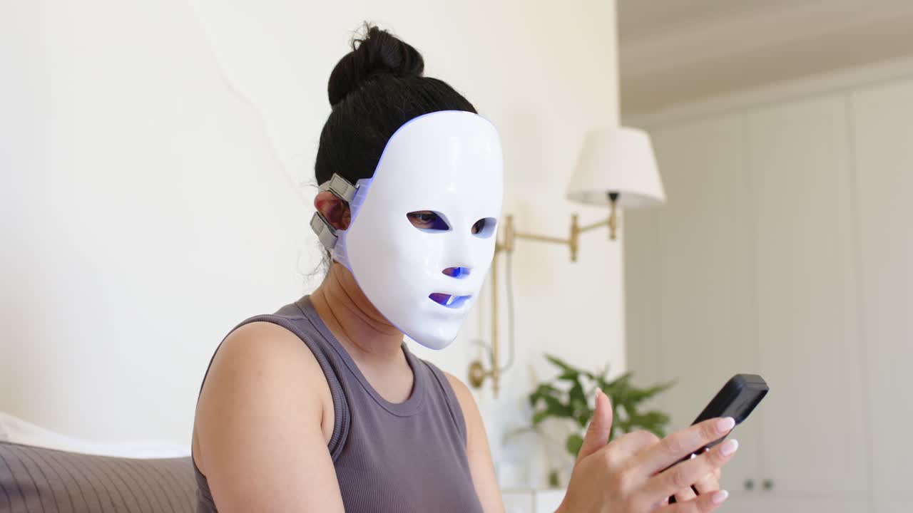 Asiatische Frau hebt Telefon, tippt auf Bildschirm und passt LED-Maske auf dem Sofa während der Heimpflege an