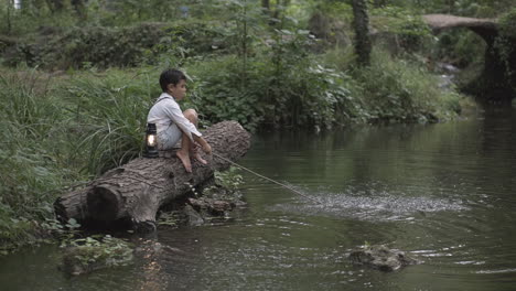 Un-Niño-Solo-En-El-Bosque-Sentado-En-El-Tronco-De-Un-árbol-Con-Una-Linterna-Y-Usando-Un-Palo-Para-Salpicar-Agua-En-El-Río