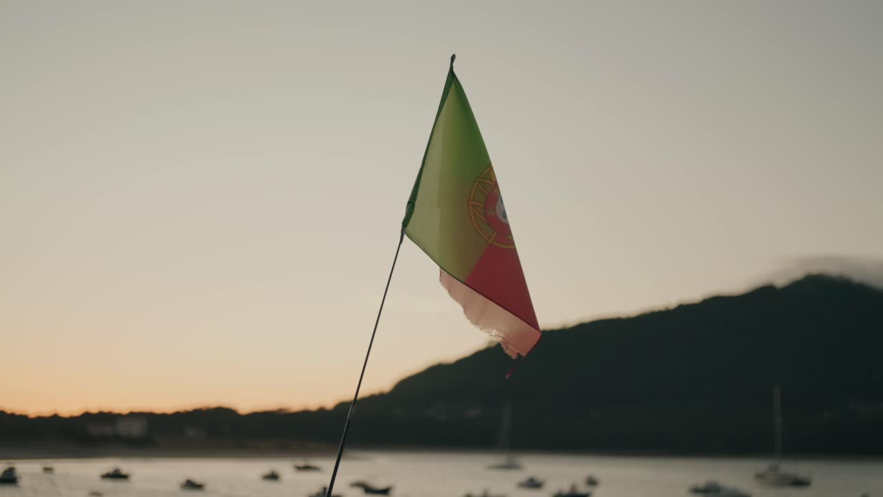 Primo piano della bandiera nazionale portoghese che sventola al vento al tramonto con montagne e barche sullo sfondo