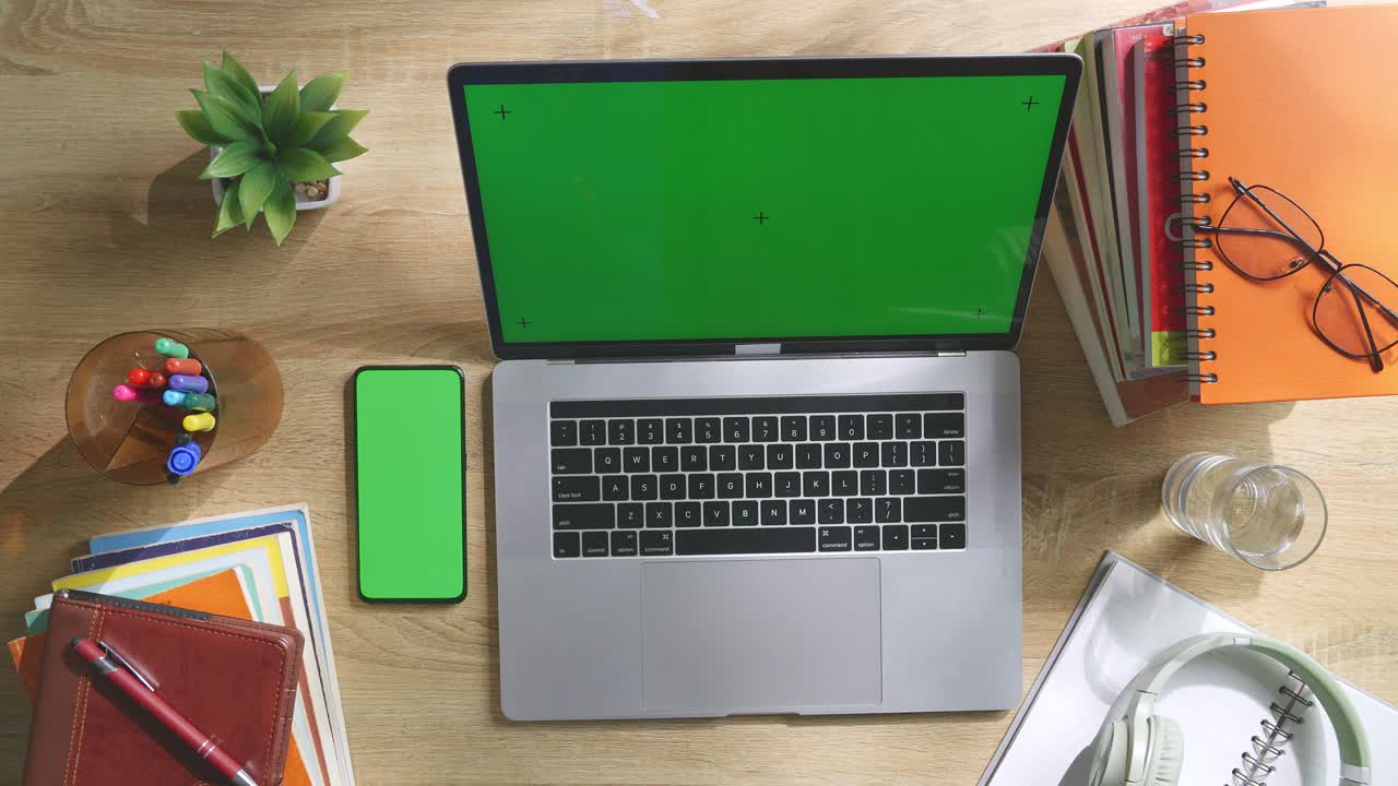 Top down view of a laptop computer with mock up green screen chromakey display on a wooden office desk next to notebook with pens, glasses, and a glass of water. Slow zoom out, close up