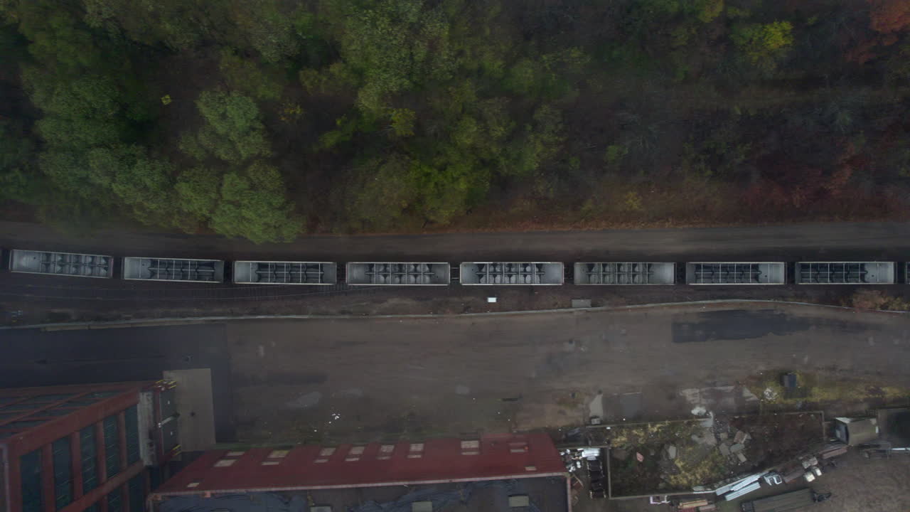 Aerial drone view tracking high overhead of a long freight train passing through an industrial area bordered by trees and scattered debris