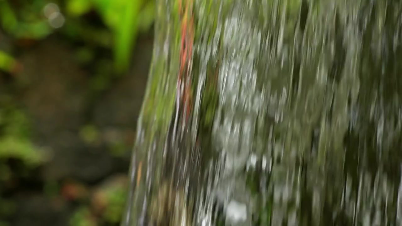 Clear fresh water flows smoothly in a continuous curtain over a ledge with blurred forest greens.