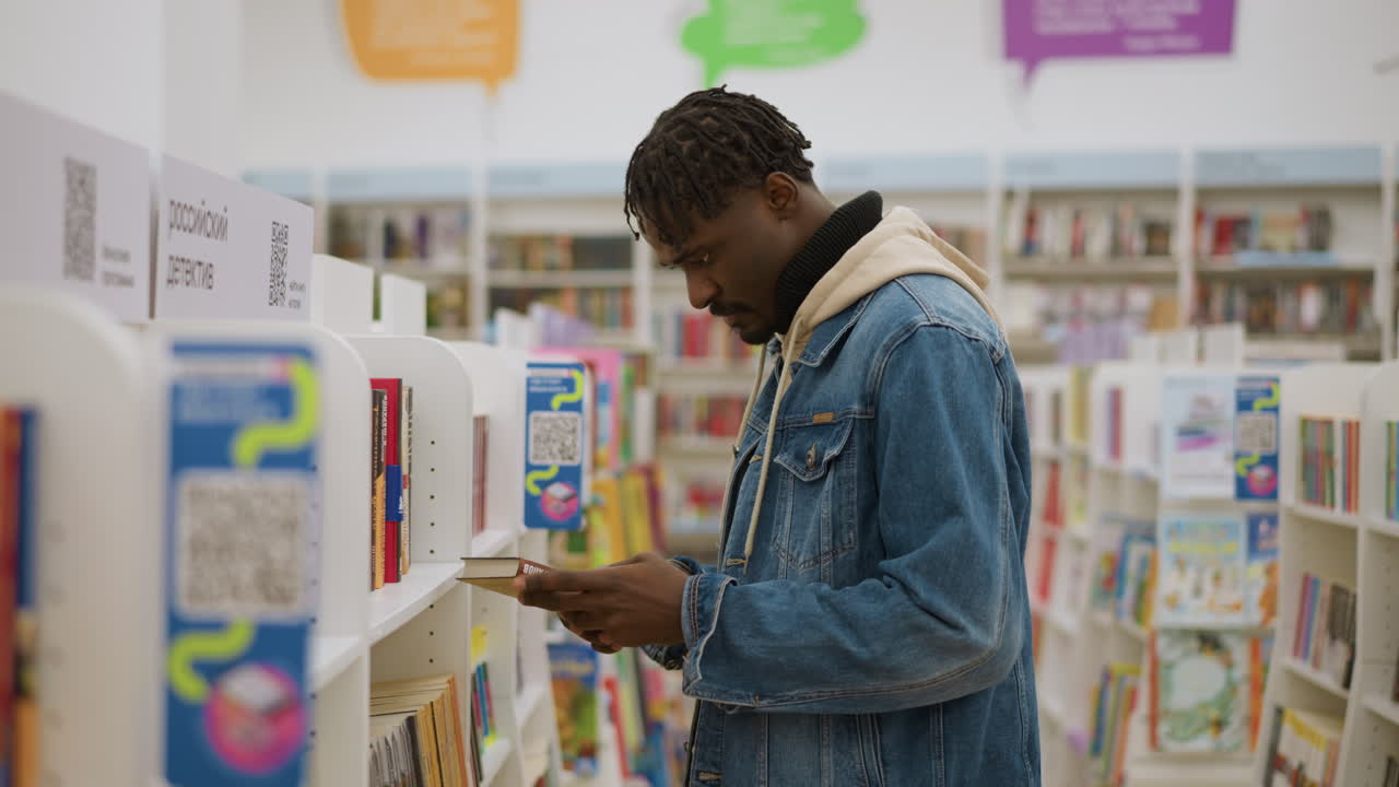 Confident young man in denim jacket standing in library, looking at bookshelves, focused on selecting books in well-organized space, embodying calm, thoughtful moment in study environment
