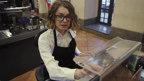 Cheerful-Saleswoman-In-White-Shirt-And-Black-Apron-Scanning-Product,-Fruits-At-Checkout-Counter-In-Bright-Supermarket-And-Putting-It-Into-Brown-Paper-Bag,-Take-An-Ec-From-Customer-And-Swipe-It