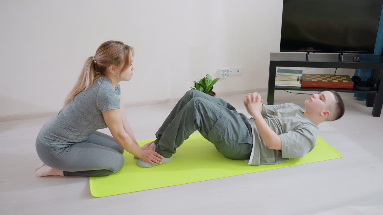 Woman supports boy practicing abdominal exercise on bright green fitness mat indoors, holding his feet as he prepares sit ups, demonstrating teamwork