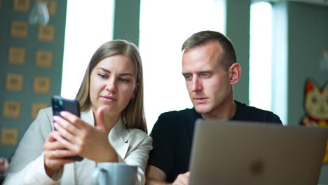 male and female sitting in the room with laptop in front of them. couple looking at the phone talking and smiling. blurred backdrop.