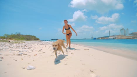 Una-Mujer-Joven-Y-Un-Perro-Corriendo-Hacia-La-Cámara-En-Una-Playa-De-Curacao