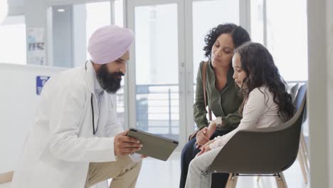 Biracial-sikh-doctor-in-turban-with-tablet-talking-to-girl-with-mother-in-hospital,-slow-motion