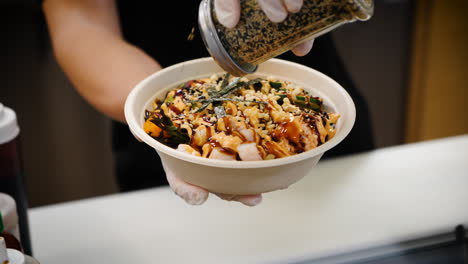 Worker-shakes-toasted-sesame-seeds-over-traditional-hawaiian-poke-salad-bowl,-slow-motion-close-up-4K