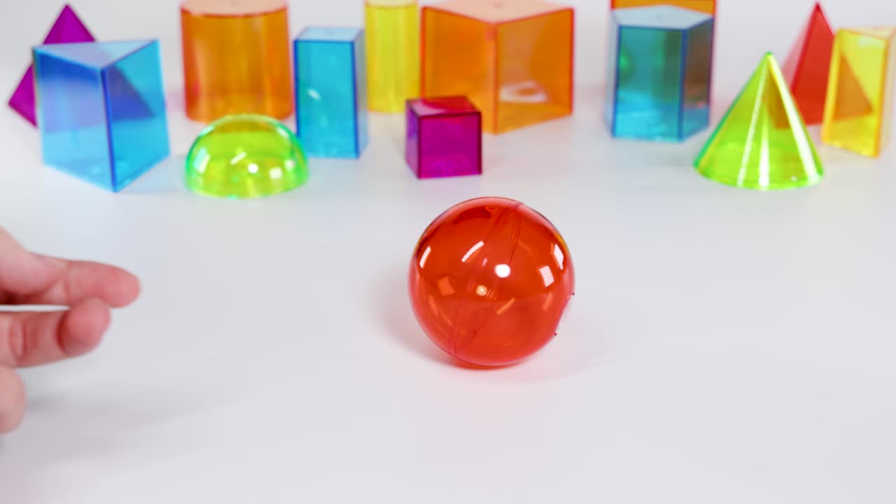 A hand interacts with colorful 3D geometric shapes, focusing on a rolling red sphere on a white surface