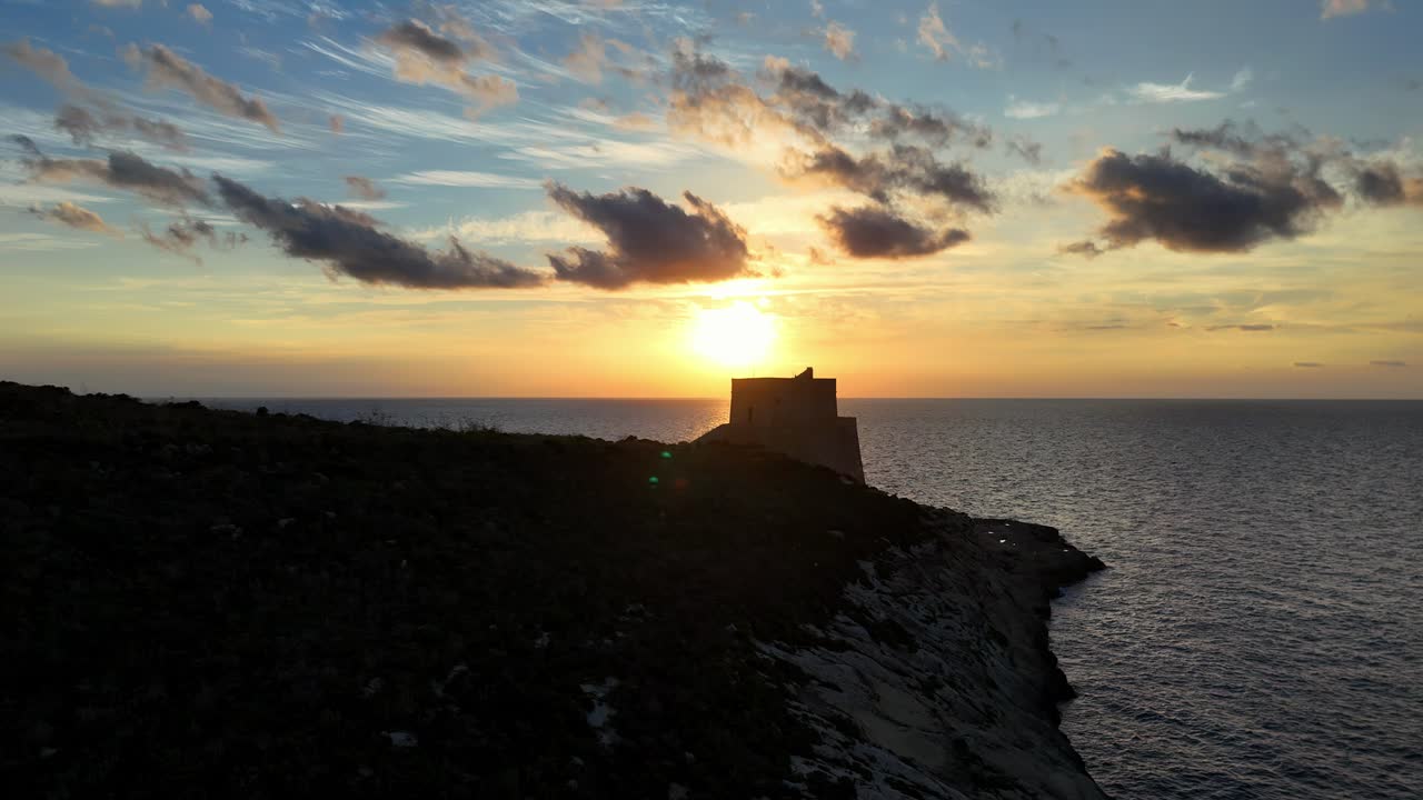 Vista da hora dourada na ilha de Gozo, apresentando uma histórica torre de vigia de pedra silhuetada contra um vibrante pôr do sol laranja, com nuvens tênues à deriva sobre as calmas e escuras águas costeiras do Mediterrâneo
