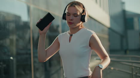 white lady in white top holding smartphone and wearing headphones dances joyfully outdoors with sunlight illuminating modern residential building and parked cars in urban setting