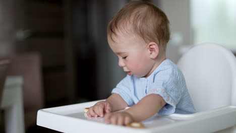 lovely baby boy sitting at the feeding table playing cheerfully with food. active toddler moving bagels by the table. blurred backdrop.