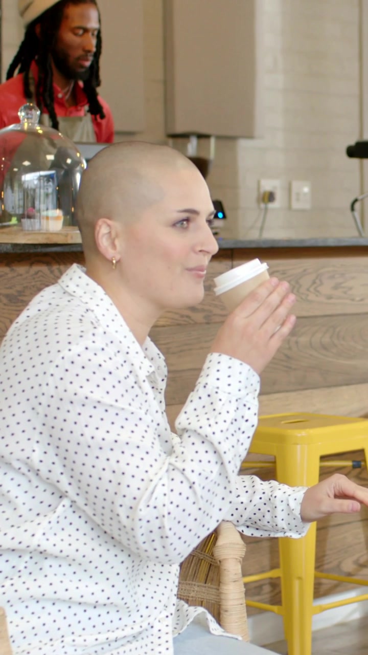 Video verticale: Una donna con la testa rasata e una camicia a pois riceve una tazza da asporto e sorseggia al bancone di un caffè.