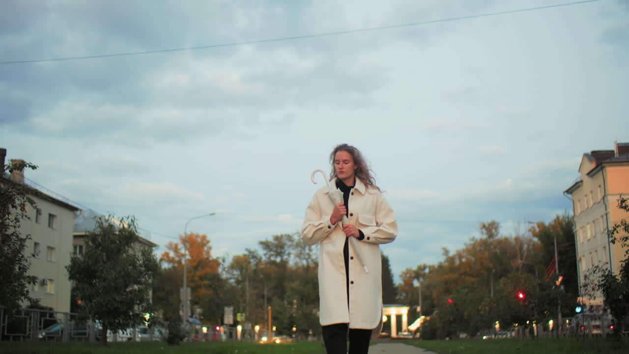 Student wearing white coat firmly holding umbrella walking along paved path surrounded by trees with blurred car lights, red traffic lights, residential buildings and glowing streetlights