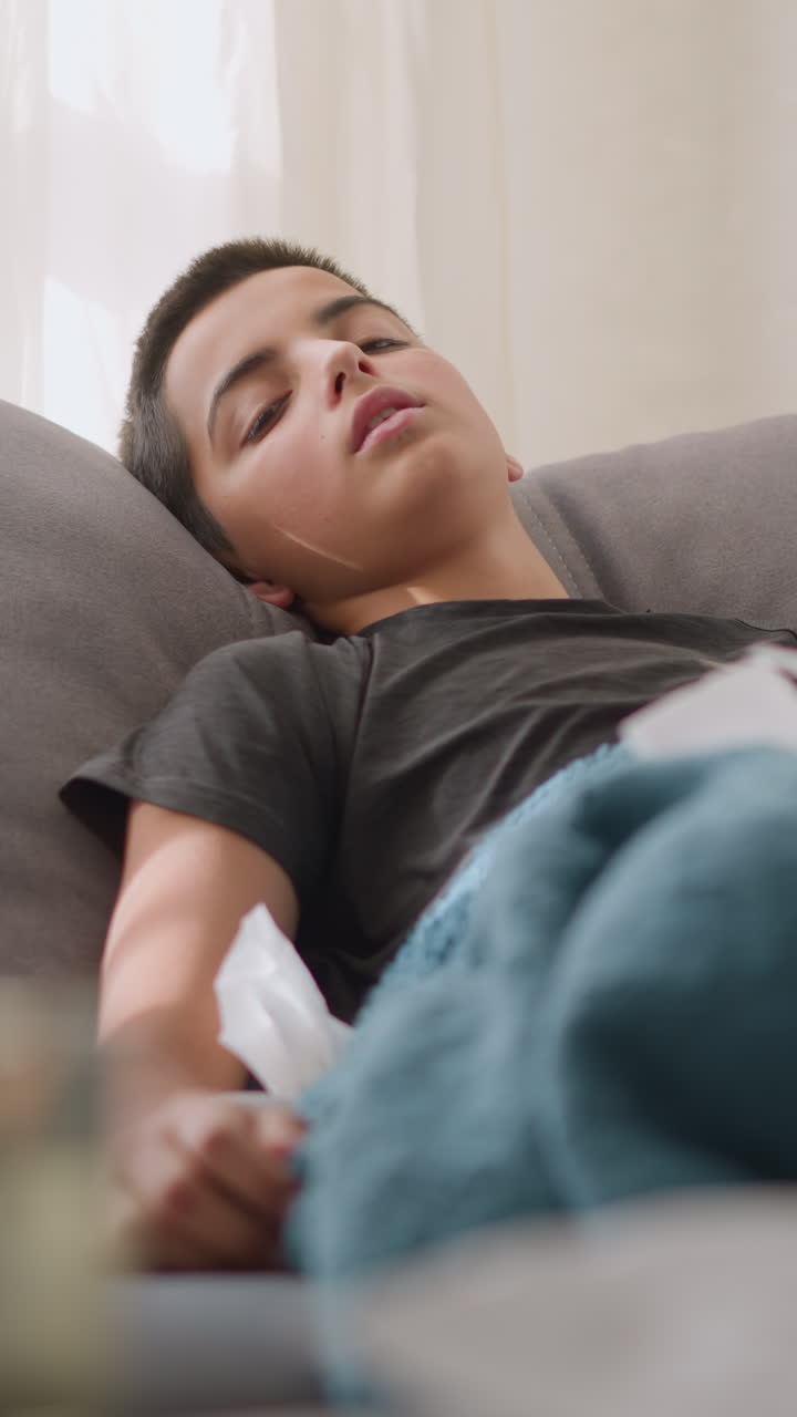 Sick child lying on the couch with low energy, surrounded by tissues, covered by a blanket, natural light coming through the window, showcasing a tired, weak child resting at home