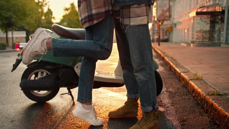 Happy-brunette-girl-with-curly-hair-in-a-plaid-shirt-and-a-white-helmet-hugs-her-boyfriend-with-curly-hair-in-a-denim-jacket-near-their-moped-on-the-street-at-dawn-in-summer