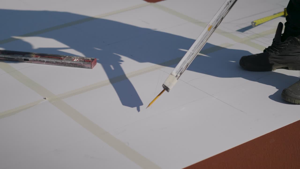 Close-up of paper sheets taped together on the ground in Neot Smadar. A man draws back and forth with a pencil, only his feet are visible - Israel
