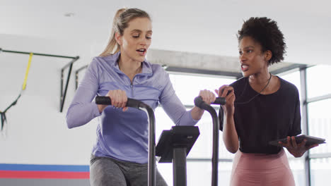 video of diverse female fitness trainer with stopwatch and woman on exercise bike working out at gym