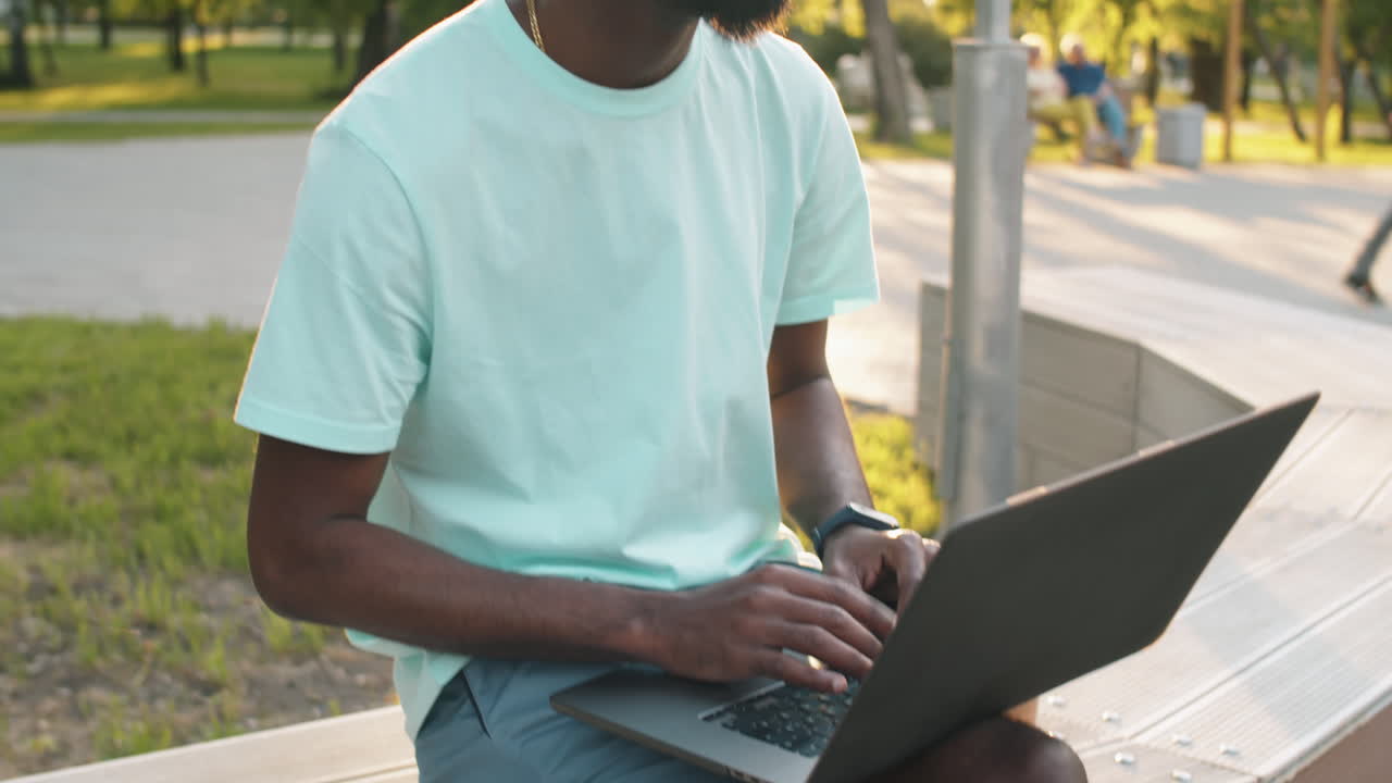 Young Black Man Sitting on Bench Outdoors and Using Laptop