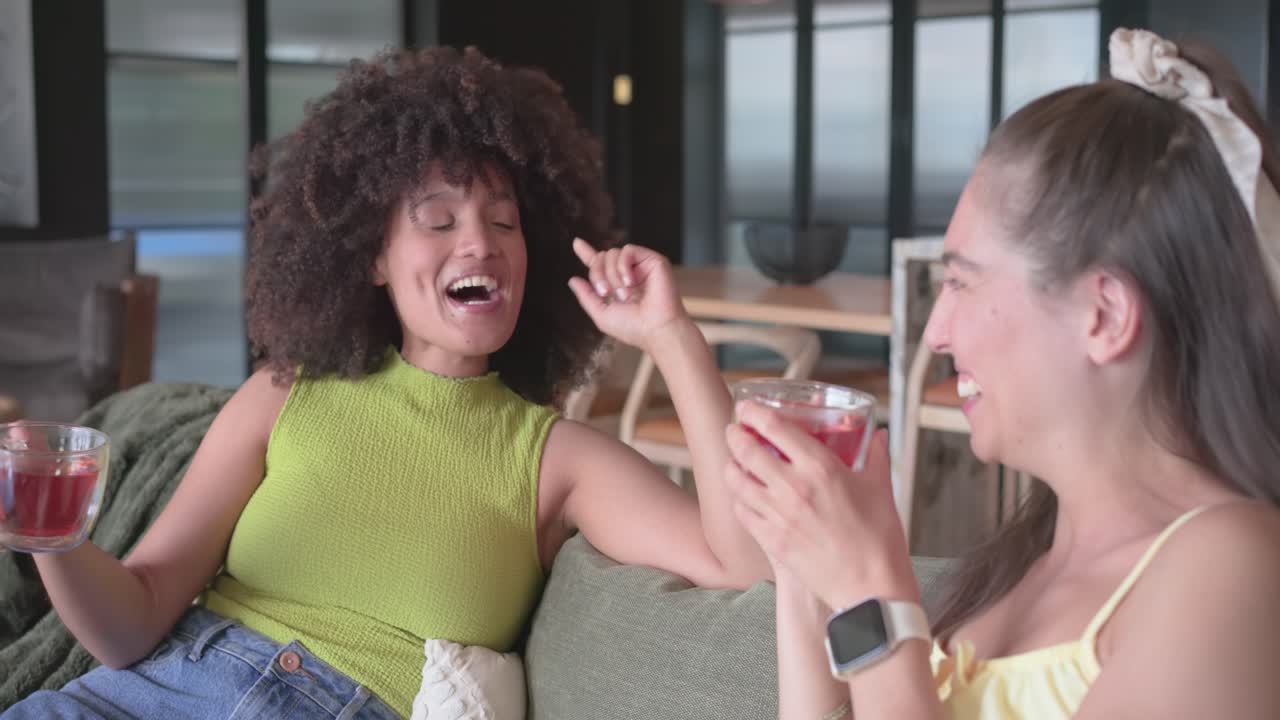 Diverse female friends sipping red tea in glass mugs on couch, woman telling story, catching up