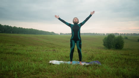 woman in green black suit stands with head slightly lifted, eyes closed, and hands slowly outstretched then brought down in a serene motion, practicing yoga in a vast open field under a cloudy sky