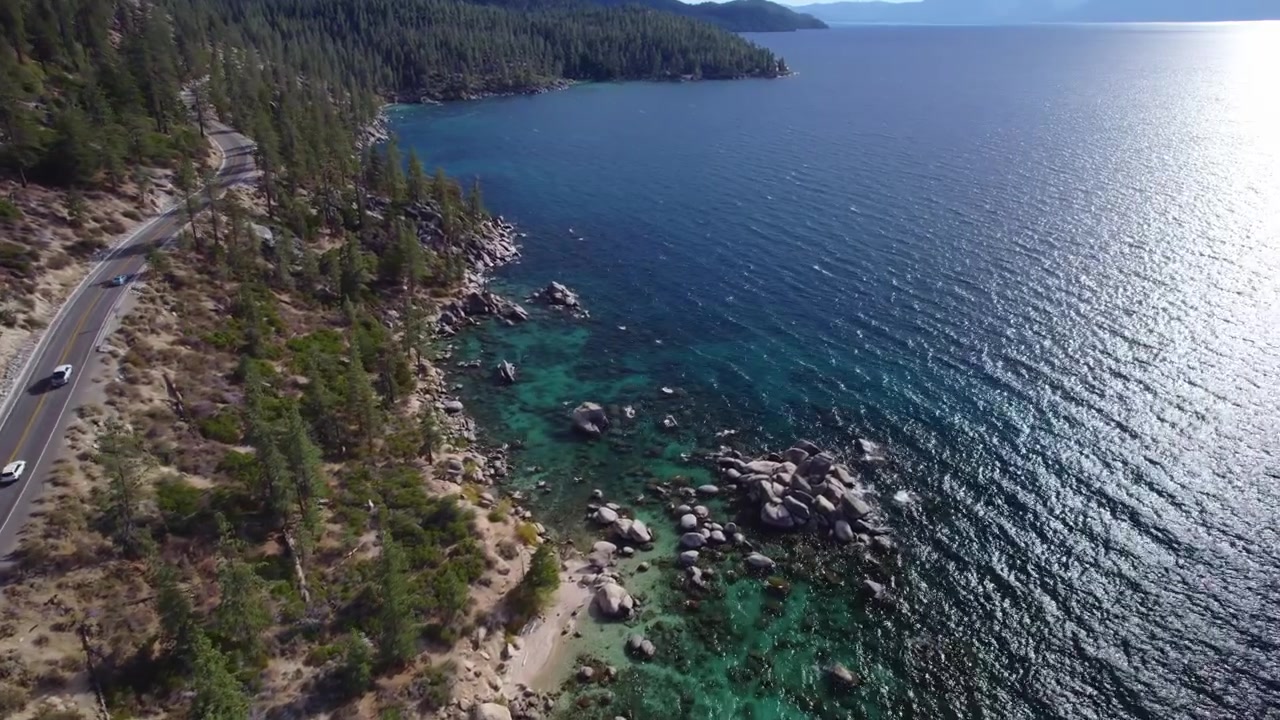 Uma tomada cinematográfica de drone em descida da costa do Lago Tahoe, apresentando uma estrada cênica com carros dirigindo ao lado de águas turquesa vibrantes
