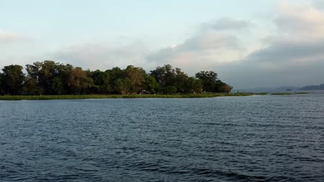 Dolly-in-aerial-drone-wide-shot-of-a-flock-of-white-egrets-flying-together-in-the-Guarapiranga-Reservoir-in-the-southern-part-of-São-Paulo,-Brazil-with-calm-waters-and-trees-during-golden-hour