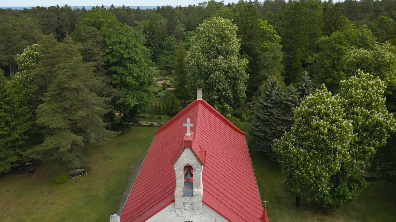 Aerial drone revealing shot showing a old country white colored church that has a red roof and bronze bell with a cross on the tower. Drone flying backwards and revealing Baltic sea in the horizon