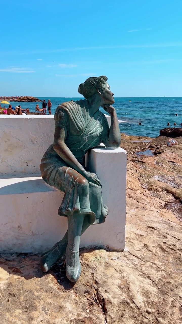 Madri, Espanha, 8 de agosto de 2025: Estátua de bronze de mulher olhando para o mar na costa. Escultura de mulher sentada em bronze em banco de concreto branco com vista para o mar Mediterrâneo em costa rochosa em Torrevieja.
