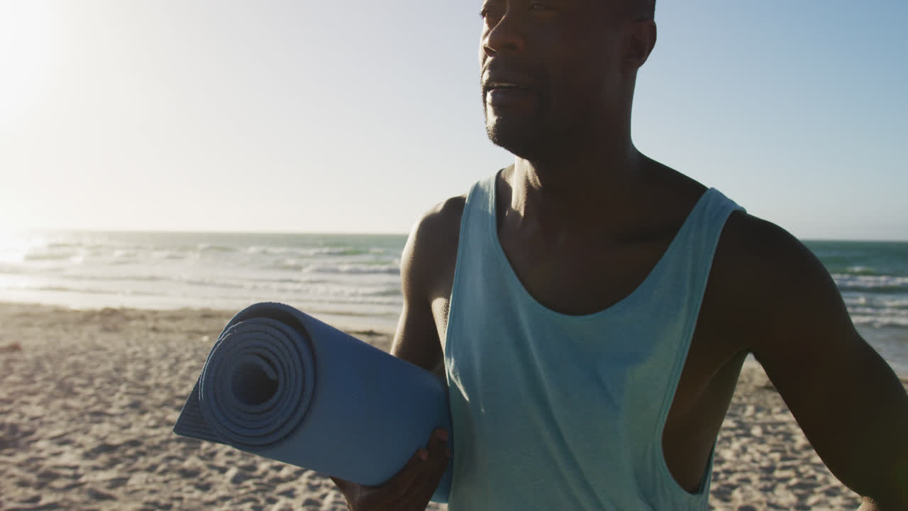Smiling african american man holding yoga mat, standing on beach by the sea