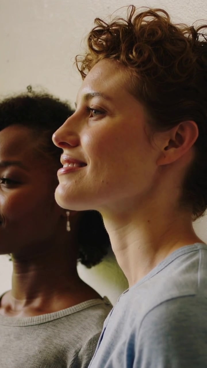 Video verticale: Donne di fronte con top grigio-blu che reagiscono a un bagliore, la telecamera si sposta verso una parete beige, polvere