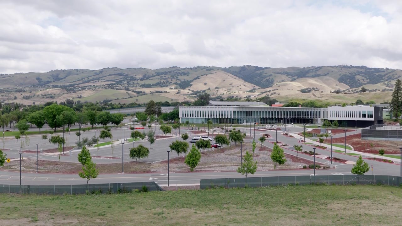 Aproximación aérea de dron hacia el campus moderno del Evergreen Valley College y su estacionamiento vacío, con las ondulantes colinas de San José y árboles al fondo, en un entorno de Silicon Valley.