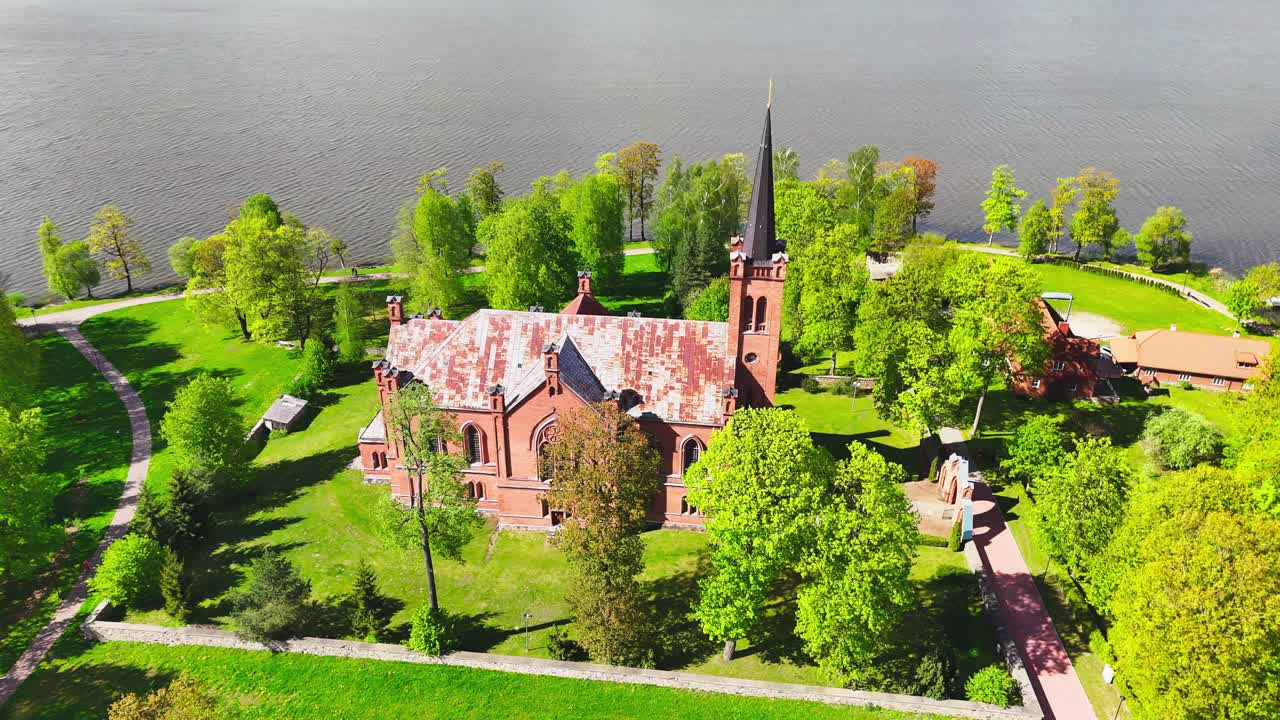 A prominent red-brick church stands by a calm river, surrounded by green trees and open lawns. The aerial view highlights peaceful spring scenery and religious architecture.