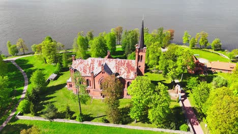 a prominent red-brick church stands by a calm river, surrounded by green trees and open lawns. the aerial view highlights peaceful spring scenery and religious architecture.