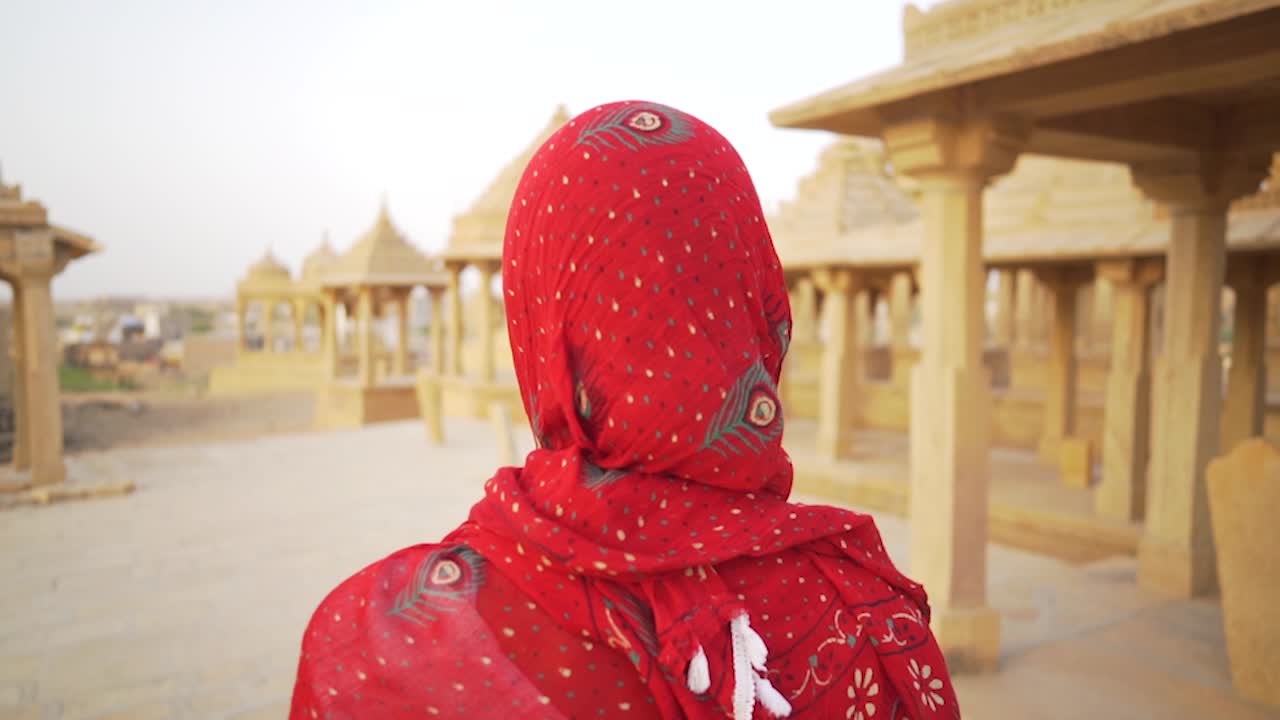A woman in a saree moves slowly and mysteriously through the sandstone cenotaphs of Bada Bagh, Jaisalmer, India, creating an atmospheric and cinematic scene.